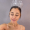 Ariana Grande aparece em frente a um fundo de cor neutra, com os olhos fechados e sorrindo levemente. Ela a coroa que sua personagem Glinda tem como acessório no filme Wicked.