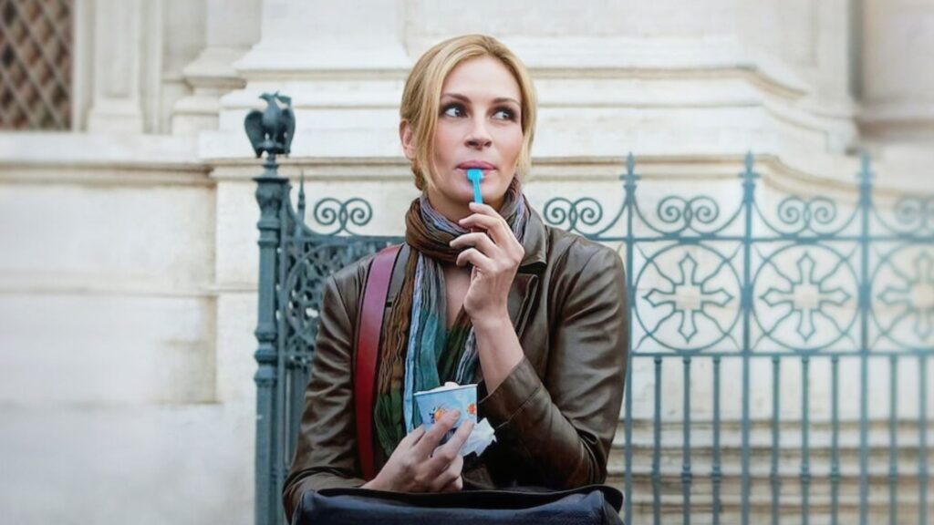 Julia Roberts, com cabelos loiros e presos para trás, sentada em frente a uma grade de ferro ornamentada com desenhos de cruzes. Ela veste um casaco de couro marrom, um lenço colorido em tons de azul e verde ao redor do pescoço e carrega uma bolsa preta sobre o colo, além de uma alça de bolsa vermelha sobre o ombro. Ela segura um copo de iogurte ou sobremesa na mão esquerda e um pequeno utensílio azul na mão direita, que leva à boca, como se estivesse prestes a comer ou provar o conteúdo. O fundo é arquitetônico, possivelmente a fachada de um prédio histórico italiano, em pedra clara. A expressão dela é contemplativa e ligeiramente sorridente, transmitindo uma sensação de leveza e tranquilidade.