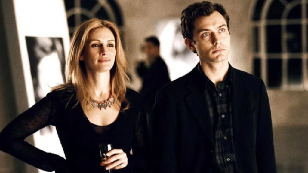 A imagem mostra uma cena do filme “Closer – Perto Demais” (2004). À esquerda está Julia Roberts, vestida com um vestido preto de mangas longas, segurando uma taça de vinho e usando um colar chamativo. Ela parece estar em um ambiente sofisticado, possivelmente uma galeria de arte, com uma expressão serena e confiante. À direita está Jude Law, usando camisa escura e blazer preto, olhando à frente com semblante pensativo. Ao fundo, a imagem está desfocada, mostrando elementos de uma galeria e outras pessoas indistintas. A iluminação é suave, destacando os rostos dos dois atores.