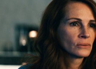 Julia Roberts está com uma expressão séria e olhar fixo, voltada ligeiramente para o lado direito. Ela tem cabelos castanhos soltos e ondulados, pele clara e veste uma blusa azul. O fundo está desfocado, com luzes suaves e tons neutros, destacando o rosto da personagem em primeiro plano. A iluminação evidencia suas feições e transmite um clima de introspecção ou tensão emocional