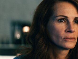 Julia Roberts está com uma expressão séria e olhar fixo, voltada ligeiramente para o lado direito. Ela tem cabelos castanhos soltos e ondulados, pele clara e veste uma blusa azul. O fundo está desfocado, com luzes suaves e tons neutros, destacando o rosto da personagem em primeiro plano. A iluminação evidencia suas feições e transmite um clima de introspecção ou tensão emocional