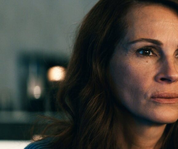Julia Roberts está com uma expressão séria e olhar fixo, voltada ligeiramente para o lado direito. Ela tem cabelos castanhos soltos e ondulados, pele clara e veste uma blusa azul. O fundo está desfocado, com luzes suaves e tons neutros, destacando o rosto da personagem em primeiro plano. A iluminação evidencia suas feições e transmite um clima de introspecção ou tensão emocional