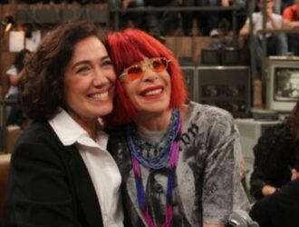 A foto mostra Lilia Cabral e a saudosa Rita Lee sorrindo, sentadas lado a lado, em um ambiente que parece ser um estúdio de televisão. Ao fundo, há uma plateia observando. Lilia veste roupa escura e Rita usa óculos coloridos e tem cabelo em tom vibrante. Ambas aparentam estar alegres, transmitindo uma sensação de amizade e descontração.