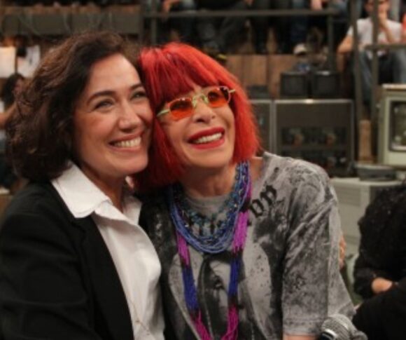 A foto mostra Lilia Cabral e a saudosa Rita Lee sorrindo, sentadas lado a lado, em um ambiente que parece ser um estúdio de televisão. Ao fundo, há uma plateia observando. Lilia veste roupa escura e Rita usa óculos coloridos e tem cabelo em tom vibrante. Ambas aparentam estar alegres, transmitindo uma sensação de amizade e descontração.