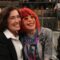 A foto mostra Lilia Cabral e a saudosa Rita Lee sorrindo, sentadas lado a lado, em um ambiente que parece ser um estúdio de televisão. Ao fundo, há uma plateia observando. Lilia veste roupa escura e Rita usa óculos coloridos e tem cabelo em tom vibrante. Ambas aparentam estar alegres, transmitindo uma sensação de amizade e descontração.