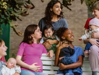 A imagem mostra cinco jovens mulheres sentadas em um banco de parque, cada uma delas segurando ou cuidando de um bebê. Elas parecem estar em um momento de convivência e troca, sugerindo laços de amizade e solidariedade. O fundo apresenta uma vegetação verde, transmitindo naturalidade e simplicidade. A foto reforça o tom humano e realista da obra, centrado na maternidade precoce e nos desafios enfrentados por essas jovens mulheres