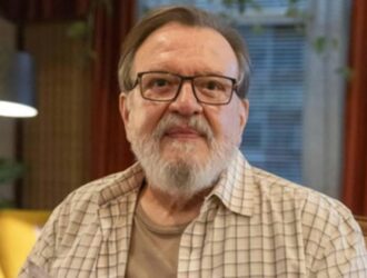 Um close-up do ator brasileiro Luis Melo, um homem mais velho, com barba e bigode grisalhos e bem aparados. Ele usa óculos de grau de aro escuro. Veste uma camisa de botões, de manga curta, com estampa xadrez em tons claros (bege, branco e marrom claro) sobre uma camiseta de cor lisa. O fundo está desfocado e apresenta tons quentes, incluindo uma cortina vermelha escura à direita e um objeto amarelo e uma luminária à esquerda, sugerindo um ambiente interno, como uma sala