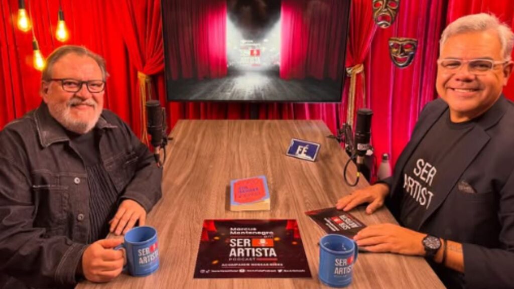 À esquerda está Luis Melo, vestindo um casaco escuro e segurando uma caneca azul.À direita está Marcus Montenegro, usando óculos, blazer preto e camiseta preta com a frase “SER ARTISTA”, também segurando uma caneca azul.Sobre a mesa, há panfletos ou livros relacionados ao tema “Ser Artista” e dois microfones de estúdio. Ao fundo, há uma televisão mostrando uma imagem com cortinas vermelhas e alguns objetos decorativos na parede, incluindo máscaras teatrais.