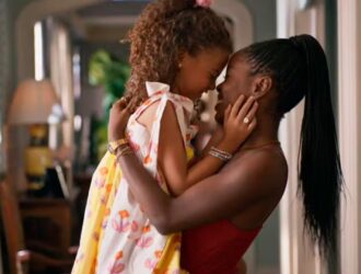 Uma mulher negra, jovem e sorridente abraça uma menina pequena e negra, encostando as testas uma na outra em gesto afetuoso. A criança veste um vestido branco com estampas coloridas e a mulher usa blusa vermelha