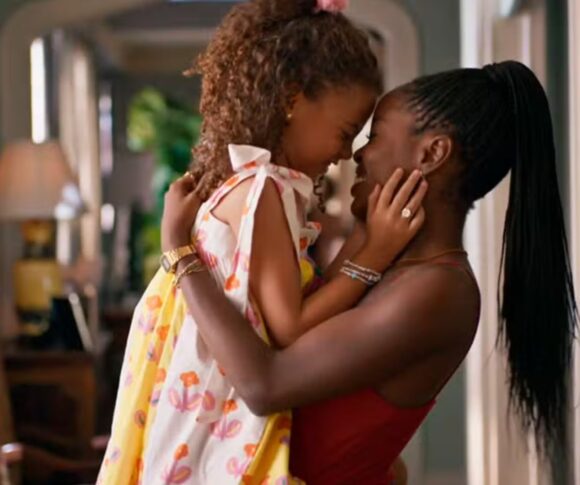 Uma mulher negra, jovem e sorridente abraça uma menina pequena e negra, encostando as testas uma na outra em gesto afetuoso. A criança veste um vestido branco com estampas coloridas e a mulher usa blusa vermelha