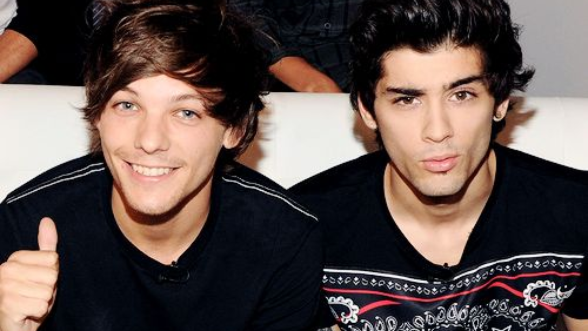 Louis Tomlinson e Zayn Malik, ex-integrantes da banda One Direction, posam juntos em um sofá. Louis sorri e mostra sinal de positivo com a mão, enquanto Zayn faz uma expressão descontraída com os lábios levemente franzidos. Ambos usam camisetas escuras.