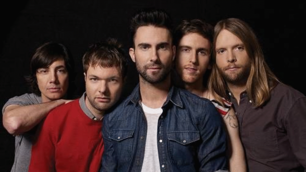 A imagem mostra os integrantes da banda Maroon 5 posando juntos, com Adam Levine ao centro, vestindo camisa jeans e camiseta branca, e os outros quatro membros ao seu redor, todos olhando para a câmera em um fundo escuro.