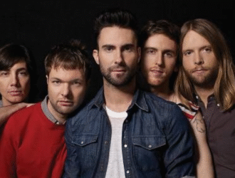 A imagem mostra os integrantes da banda Maroon 5 posando juntos, com Adam Levine ao centro, vestindo camisa jeans e camiseta branca, e os outros quatro membros ao seu redor, todos olhando para a câmera em um fundo escuro