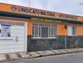 A imagem mostra a fachada de um prédio identificado como "Sindicato Nacional dos Aposentados", organização suspeita de envolvimento no esquema de desvio de pensões dos aposentadas no Brasil. A estrutura tem uma fachada nas cores laranja e branca, com calçada de ladrilhos em frente.