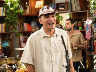 João Gomes sorri enquanto canta durante a gravação do Tiny Desk Brasil, cercado por músicos e instrumentos em um ambiente acolhedor, com estantes de livros e plantas ao fundo