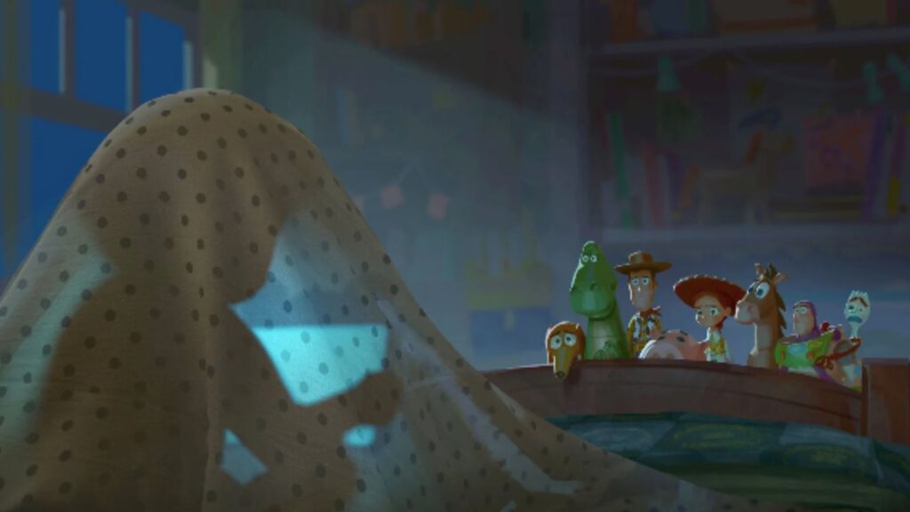 Cena noturna em um quarto infantil, iluminado pela luz azulada de uma tela. Uma criança aparece coberta por um lençol de bolinhas, segurando um tablet que brilha no escuro. À direita, os brinquedos de Toy Story — incluindo Woody, Jessie, Buzz Lightyear, Rex, Porquinho, Garfinho e outros — observam atentos e com expressões preocupadas. A imagem transmite a ideia central do novo filme, mostrando o contraste entre o encanto dos brinquedos tradicionais e o fascínio das crianças pela tecnologia.