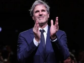 A imagem mostra o cineasta Walter Salles de pé, vestindo um terno azul-escuro com camisa branca e gravata preta. Ele está sorrindo e aplaudindo, em um ambiente que parece ser uma cerimônia ou evento formal. Ao fundo, é possível ver uma plateia desfocada, também aplaudindo, o que sugere um momento de reconhecimento ou homenagem. A iluminação destaca o diretor em primeiro plano, transmitindo uma atmosfera de celebração e emoção