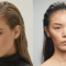 Duas mulheres posam com o cabelo no estilo wet hair, penteado com aparência molhada e polida. À esquerda, uma mulher com cabelo loiro puxado para trás com efeito molhado e pele clara usa uma blusa preta de gola alta, com maquiagem leve que destaca os olhos azuis e os lábios rosados. À direita, uma mulher com cabelo preto igualmente penteado para trás com efeito molhado exibe uma maquiagem minimalista, com foco na pele natural e sobrancelhas definidas. Ambas transmitem uma estética moderna e sofisticada